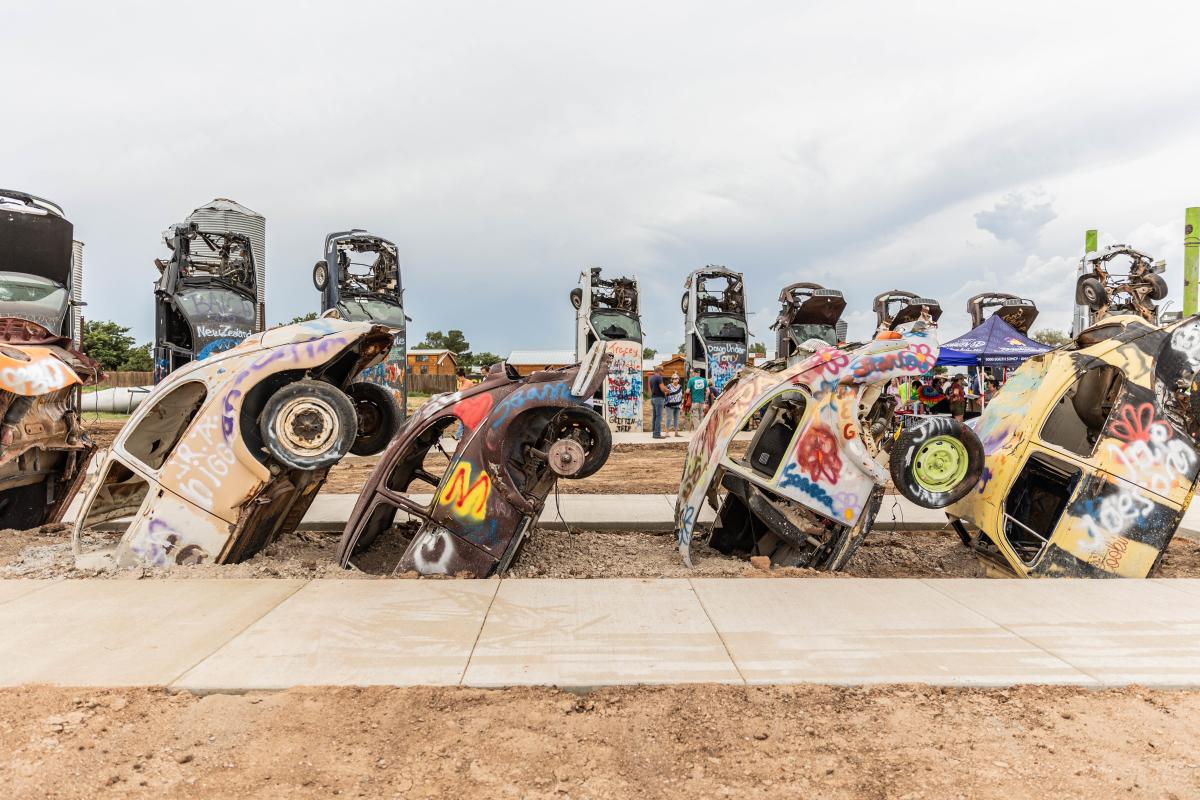 A colorful photo of Slug Bug Ranch at its new home in Starlight Ranch, Amarillo, TX. The image features several partially buried VW Beetles covered in vibrant graffiti, similar to Cadillac Ranch. Visitors are seen in the background, engaging with the art installation. This quirky and interactive attraction continues to delight with its unique blend of automotive history and creative expression.