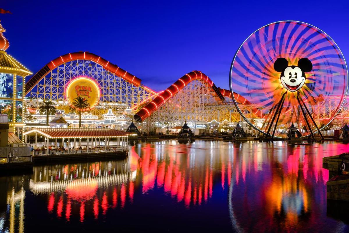 Image of Pixar Pier at Disney California Adventure Park lit up at nighttime. Pixar Pier Pal-A-Round can be seen to the right of the image. To the left of the image, a roller coaster, the Incredicoaster, can be seen. Both attractions overlook a large 'lagoon' of water.