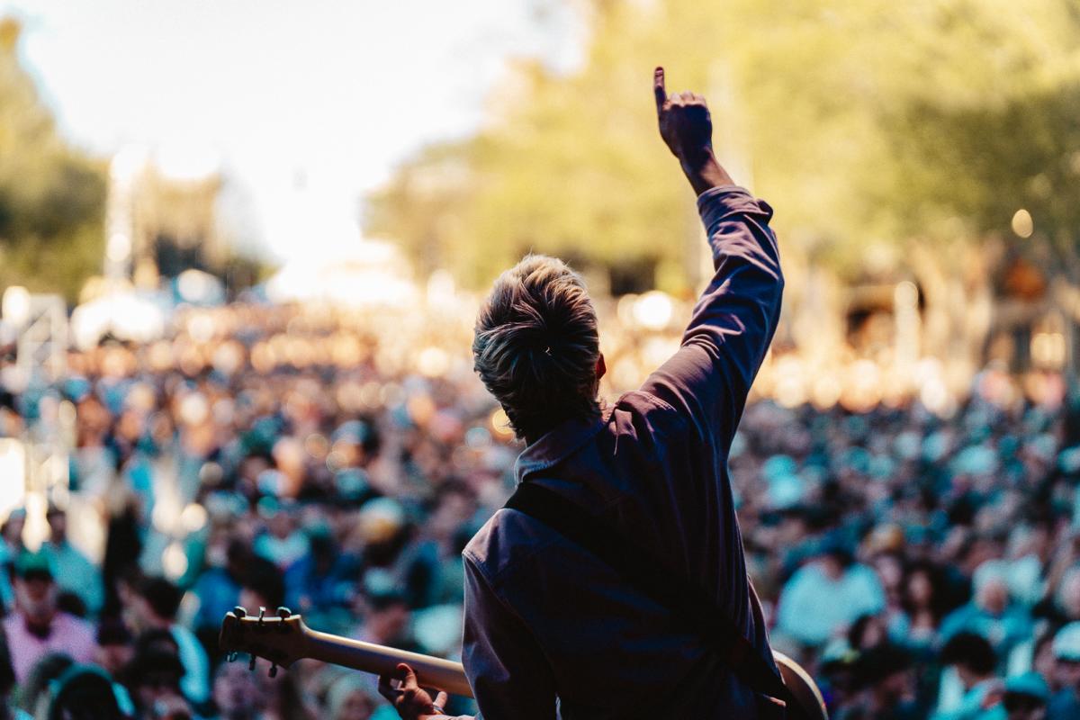 A performer stands with his back to the camera, holding a guitar, and a large crowd can be seen in front of him.