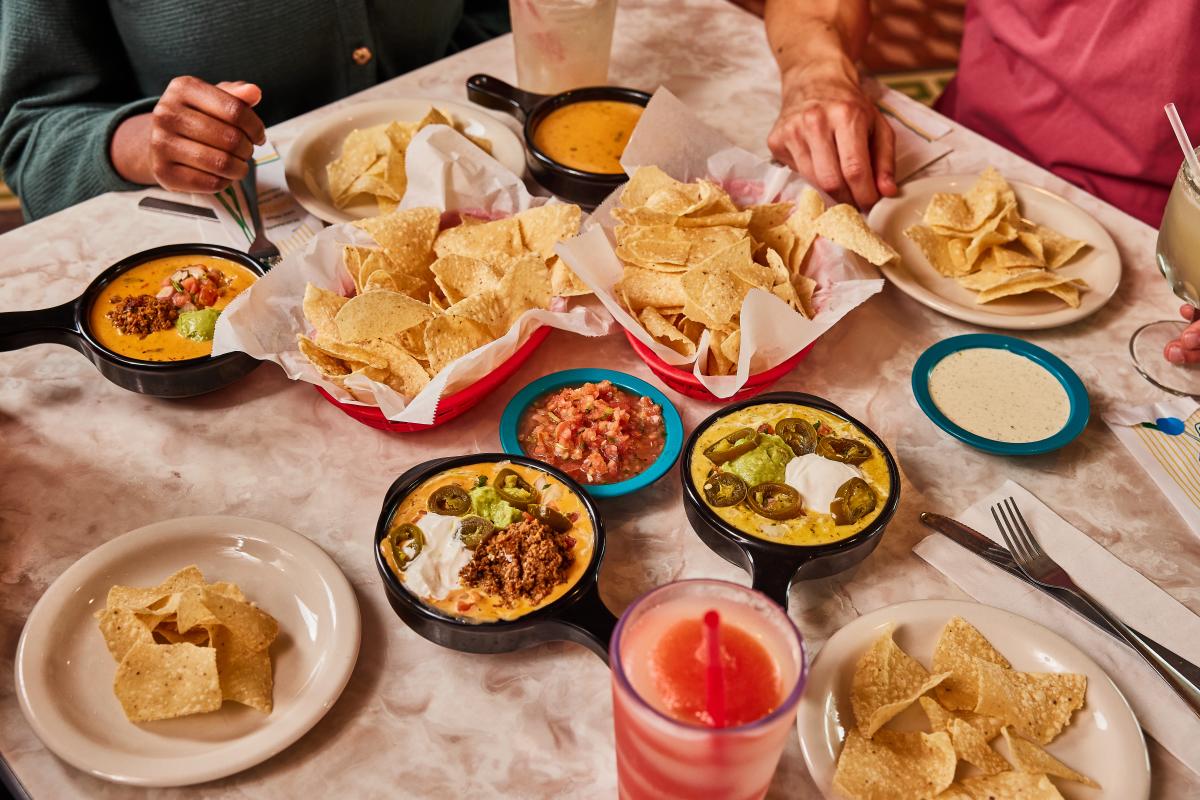 A table filled with a basket of chips and four different queso dishes from Chuy's along with salsa and other Tex-Mex.