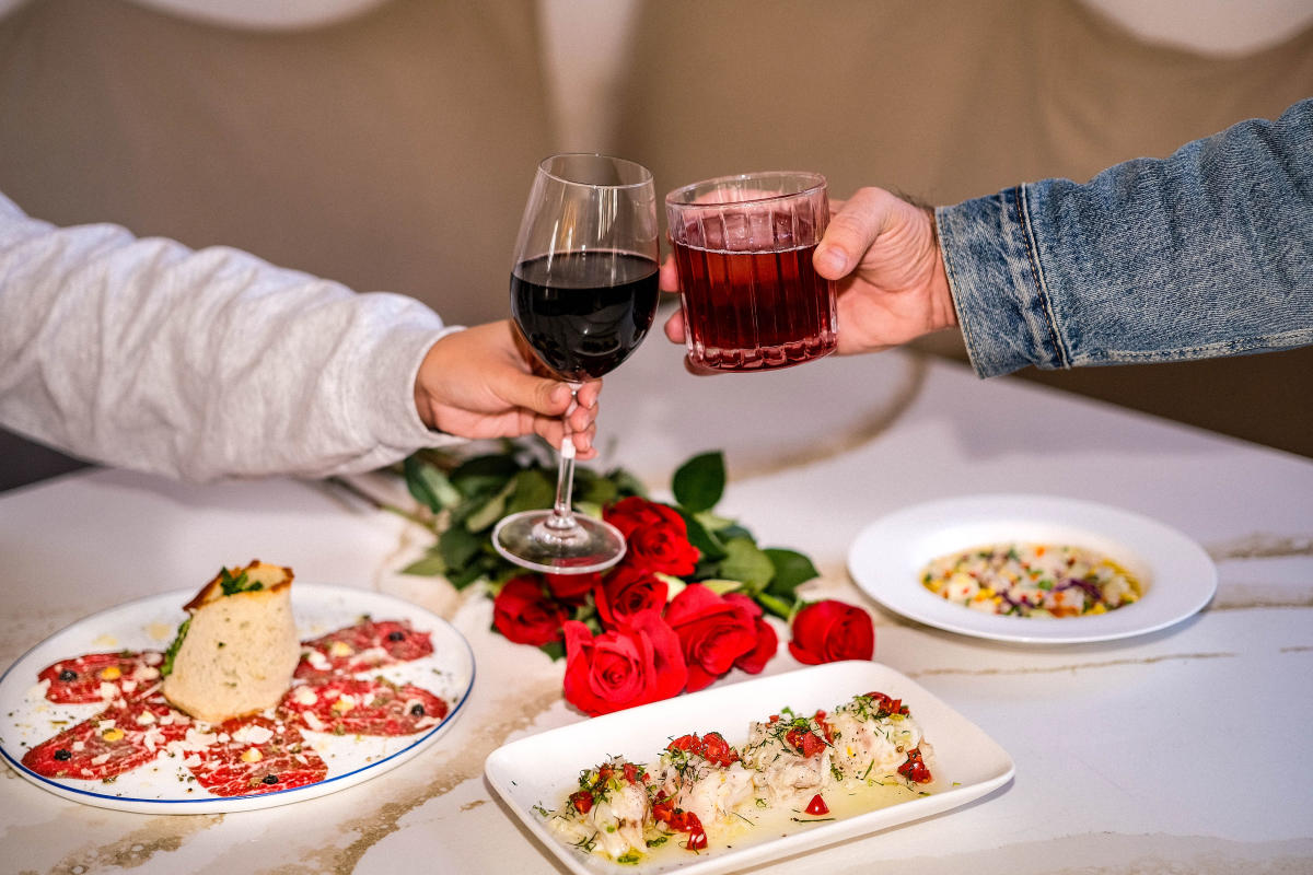 Close up image of three different dishes sitting on a table with a bouquet of roses and two people toasting with glasses.