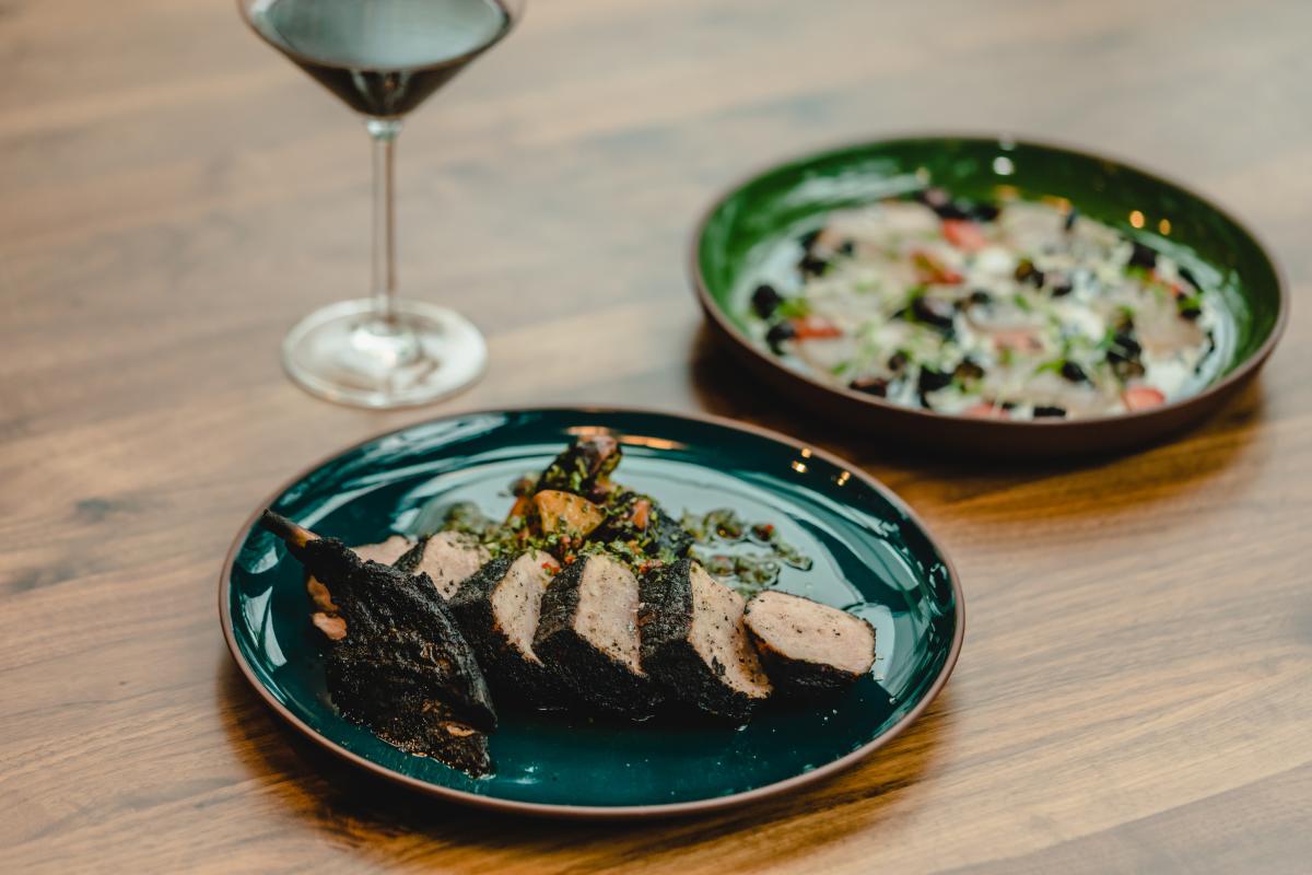 Image of a meat meal on a blue plate with a wine glass and additional plate on the table in the background.