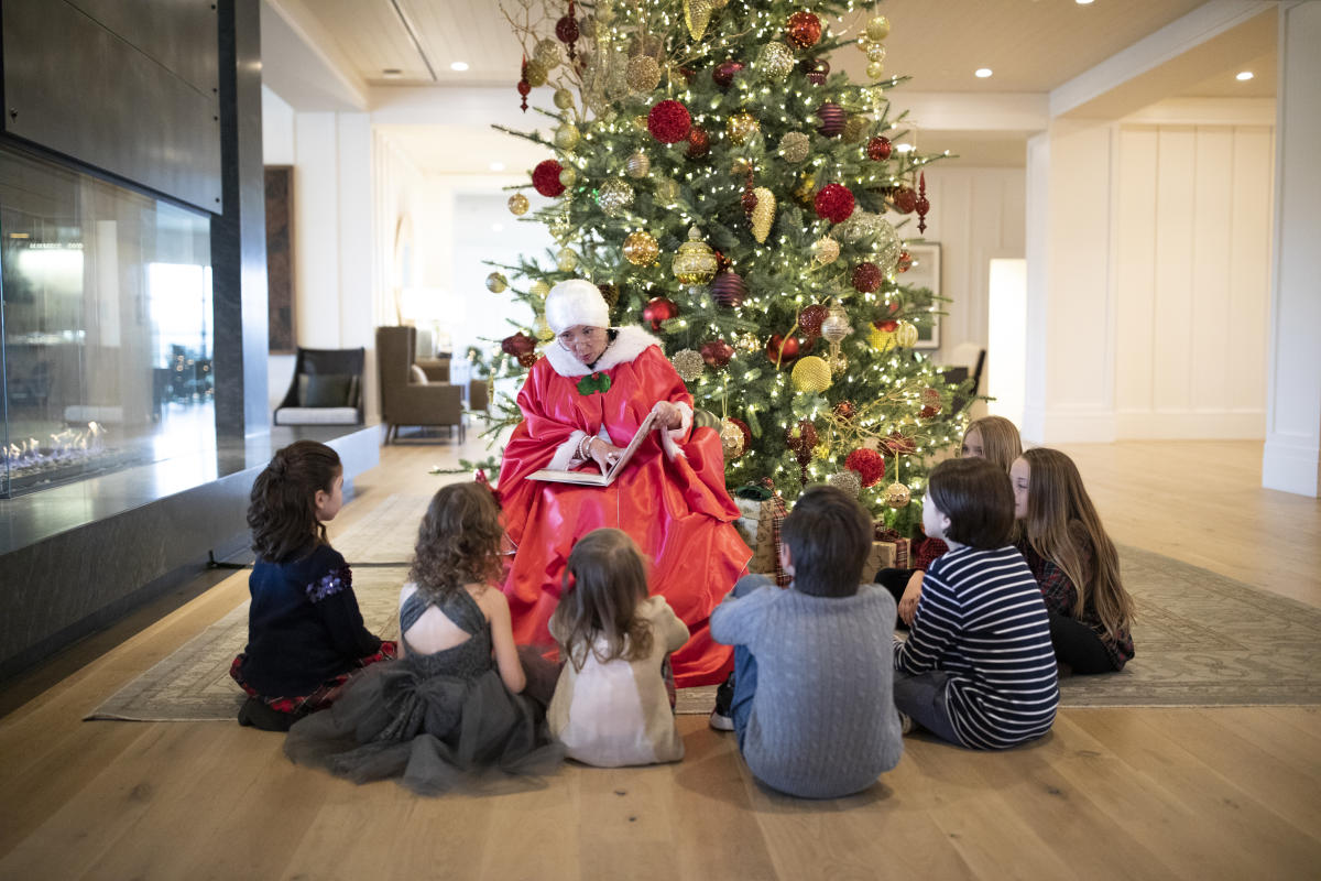 Mrs. Claus sitting in front of a Christmas Tree reading a story to a group of young kids sitting in front of her in a circle.