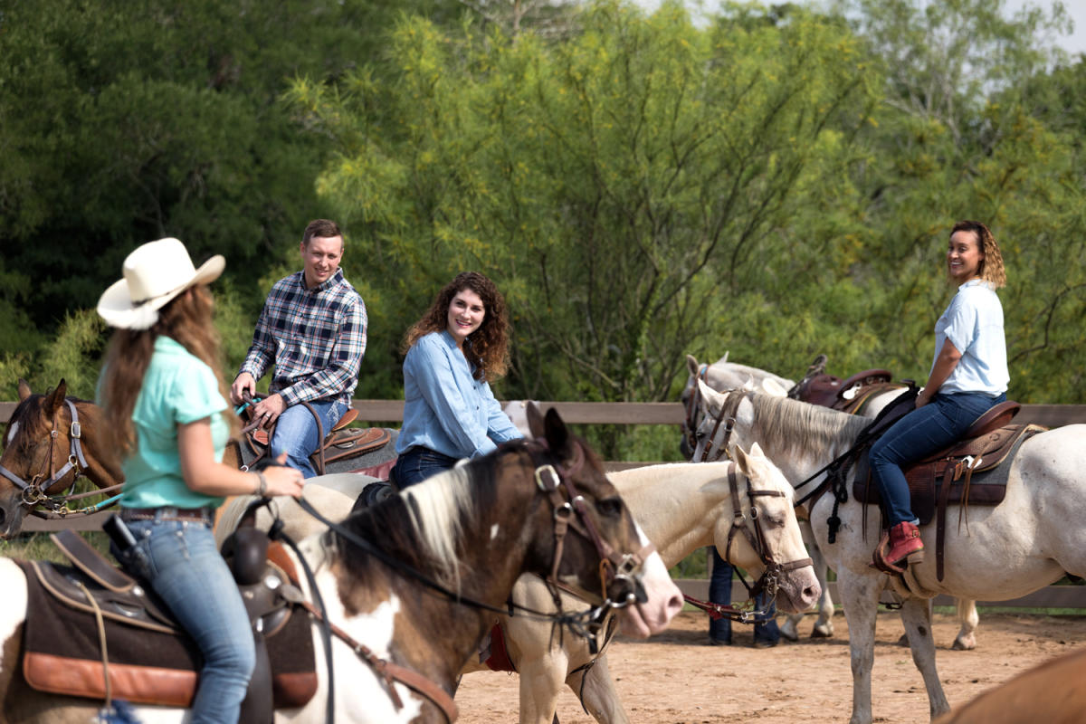 Image of four people on horseback riding in a corral with green trees in the background.