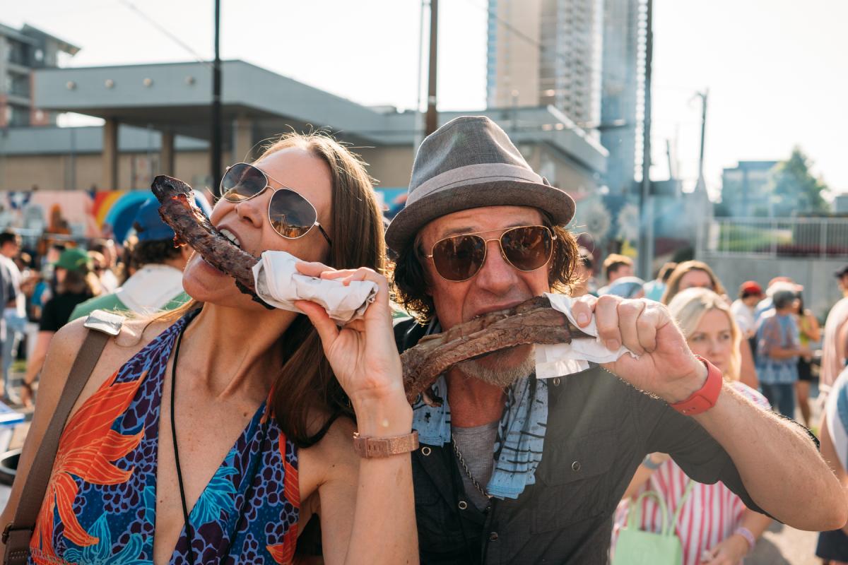 Image of a man and woman holding up a large beef rib to their mouths and taking a bite.