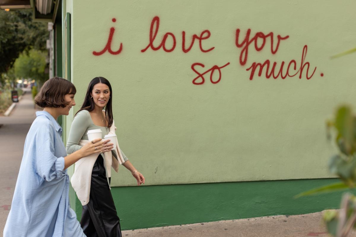 Two women walking in front of the I Love You So Much mural with coffee in hand.