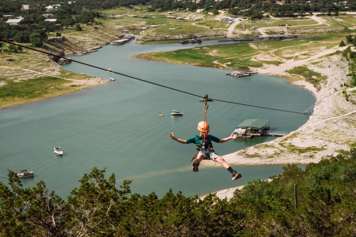 A young boy going on a zipline with Lake Travis below him in the background.