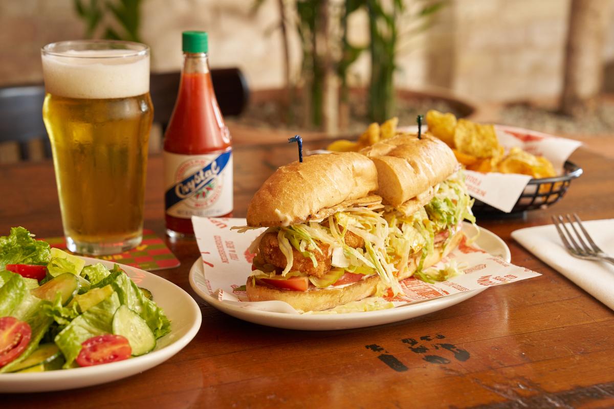 A shrimp po-boy surrounded by a salad, draft beer and hot sauce all sitting on a table at Uptown Sports Club.