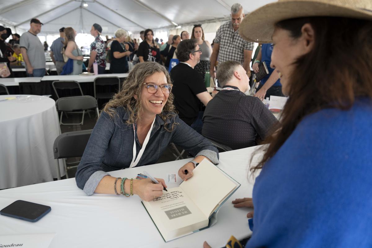 Image of a women smiling up a women from a table while she signs a book.