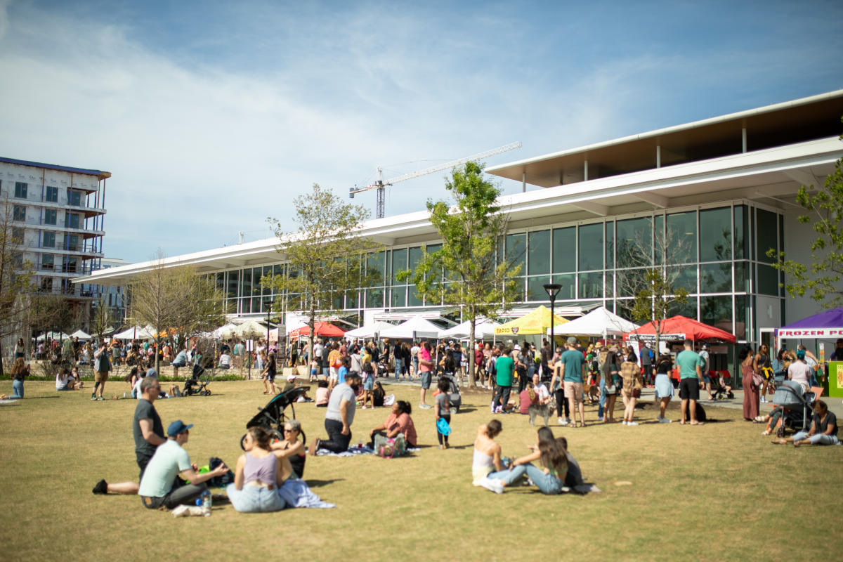 Mueller park and farmer's market tents, with people enjoying their market finds on picnic blankets in the grass.
