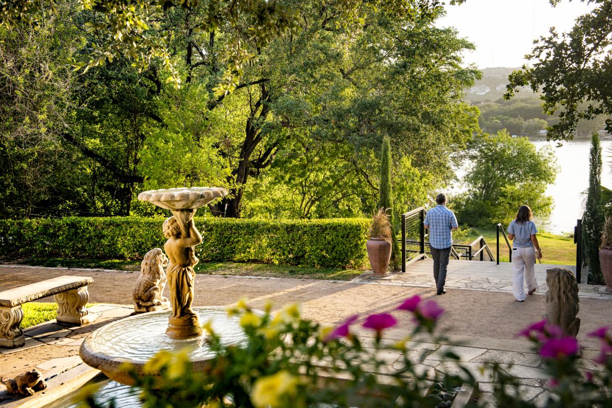 Image of a man and a woman walking down the outdoor steps towards Lake Austin with a water feature and statue in the foreground at The Contemporary Laguna Gloria.