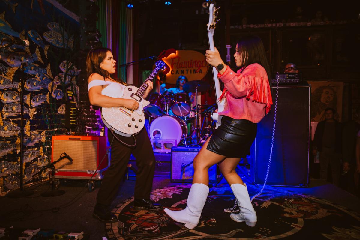 Two Latina women playing guitars on a stage with colorful lights.