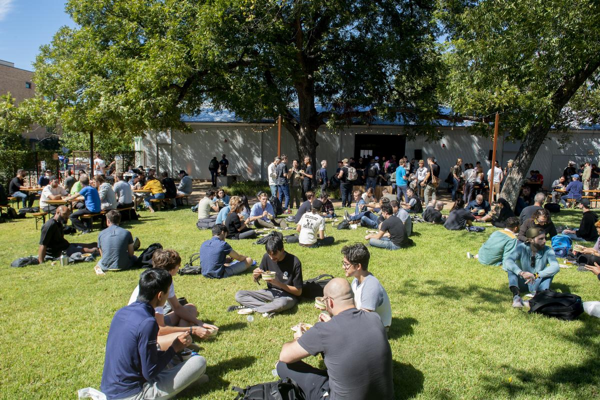 Large group of people sitting on the outdoor greenspace at Distribution hall with trees and string lights in the background.