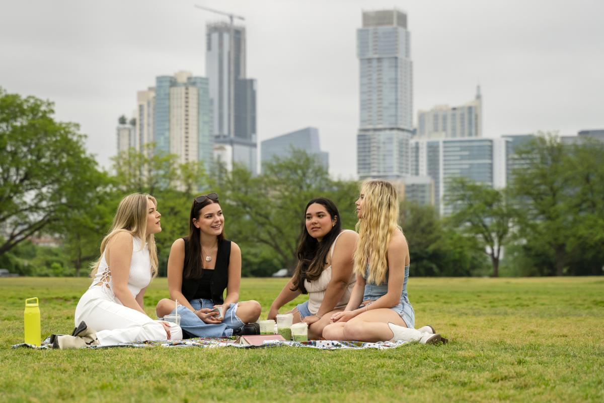 Image of four women sitting on a blanket in Zilker Park chatting with the Austin skyline in the background.