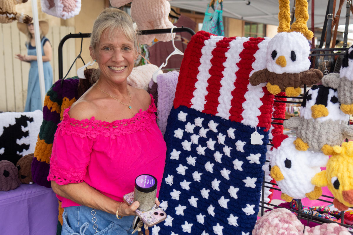 Woman with blonde hair in a pink top posing for a picture next to an crocheted American flag blanket.