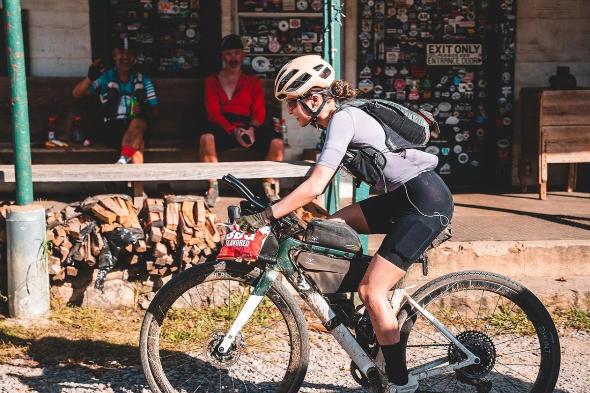 A cyclist in a helmet rides a bike near a rustic building adorned with stickers. Two people relax on a porch in the background.