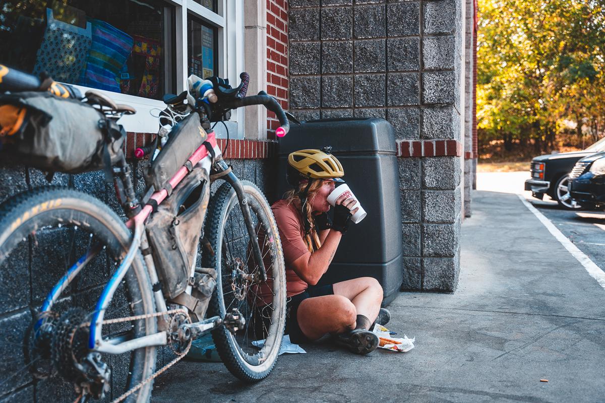 A cyclist in a helmet sits on the ground, sipping coffee beside a parked bike. A store entrance and cars are visible in the background.