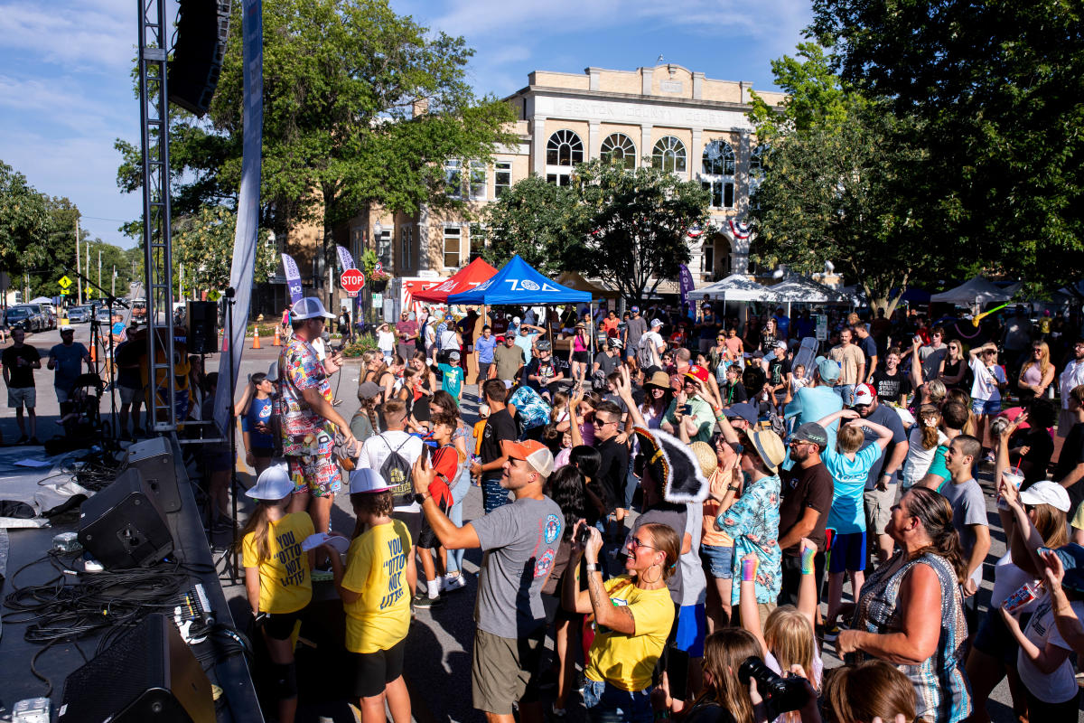 First Friday with a large crowd enjoying a performance. Colorful tents and a stage are visible, surrounded by trees and historic buildings.