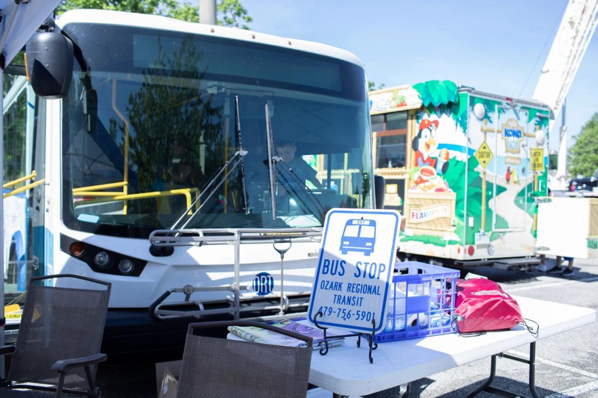 Ozark Regional Transit bus at a designated stop with a 'BUS STOP' sign displaying contact information, set against a sunny backdrop with a colorful ice cream truck in the distance.