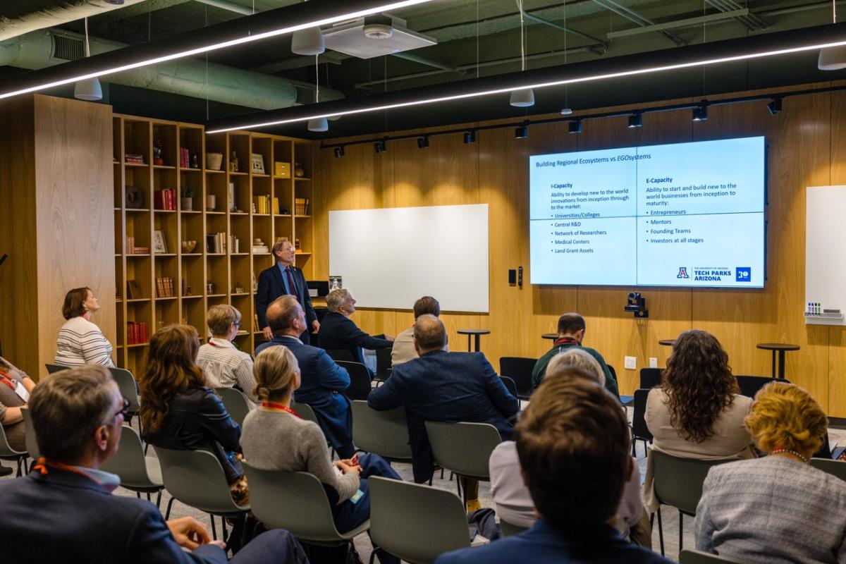A speaker presents in a modern conference room with a large screen displaying information. Attendees sit in rows, engaged in the session.