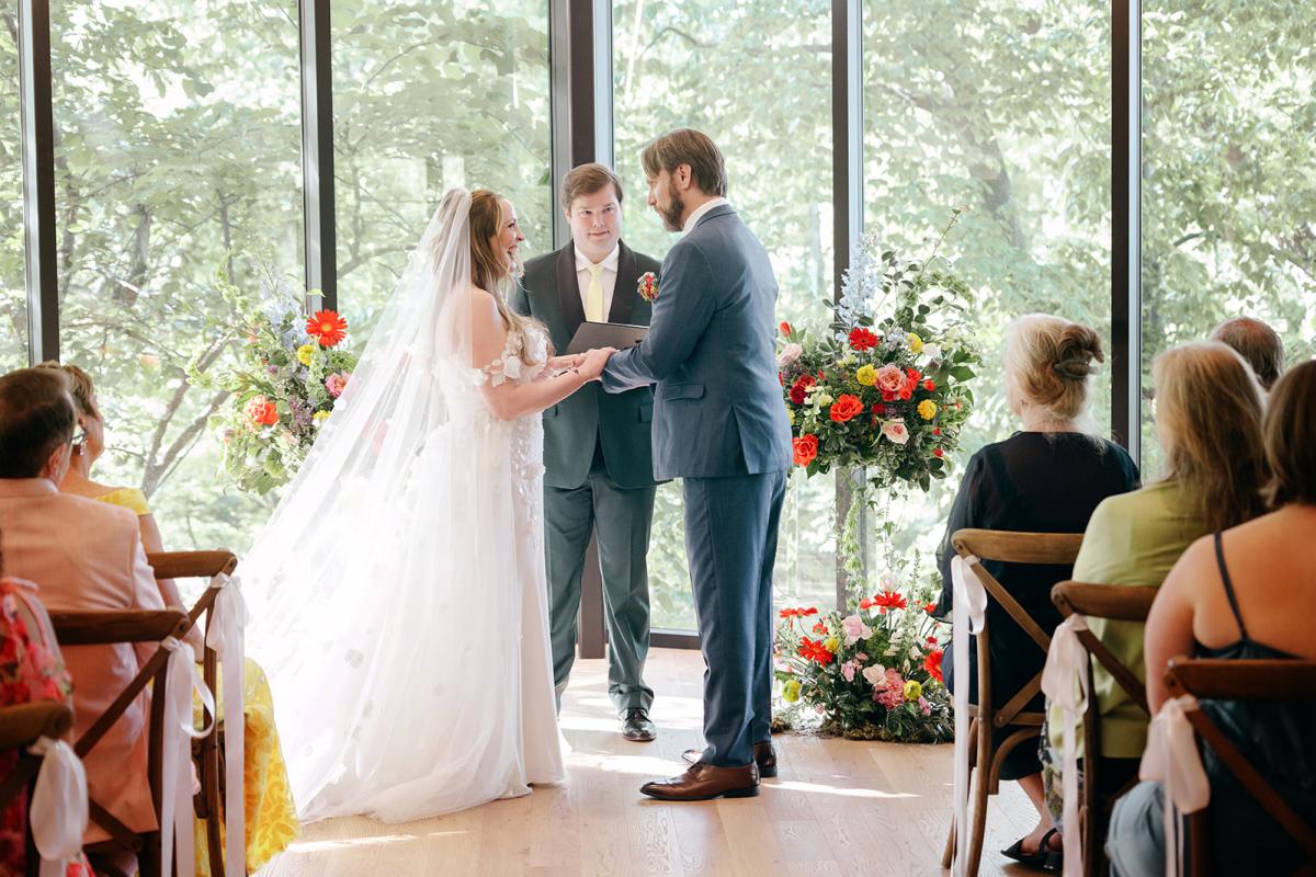 A bride and groom stand facing each other holding hands during a wedding ceremony, with an officiant and guests seated around them in a room with large windows and floral arrangements.