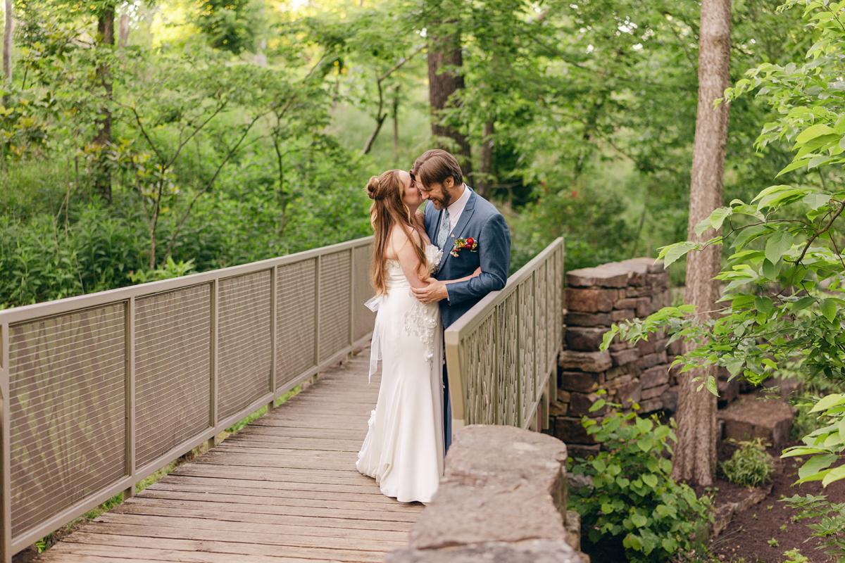Bride and groom kiss while standing on a wooden footbridge surrounded by trees and greenery.