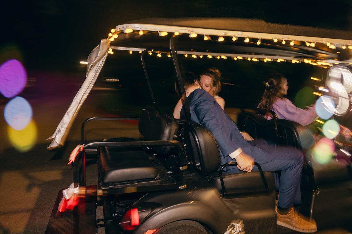 A newlywed couple shares a kiss while riding in a decorated golf cart at night, with string lights illuminating the vehicle and colorful lights creating a blurred background.