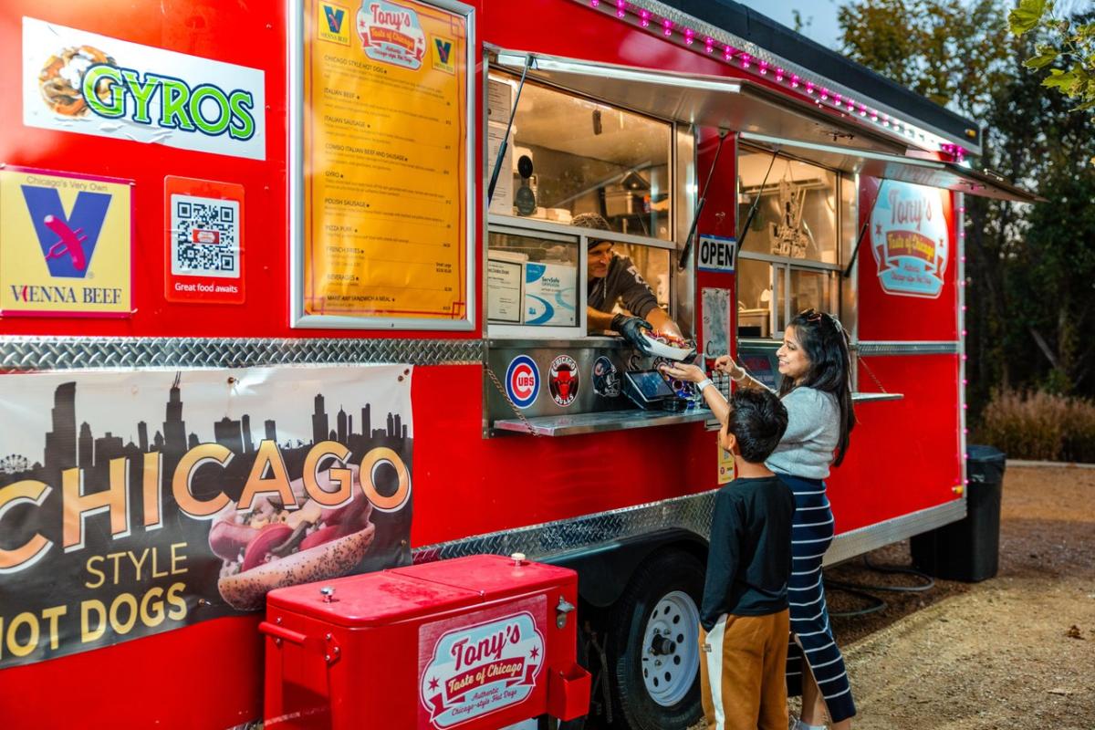 A food truck with a red exterior serves Chicago-style hot dogs. A woman and child order from the window, surrounded by city-themed decor.