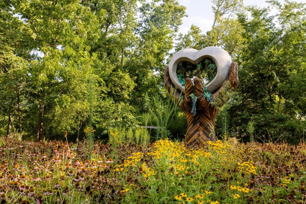Sculpture of two hands holding a heart-shaped frame, surrounded by wildflowers and greenery at Compton Gardens.