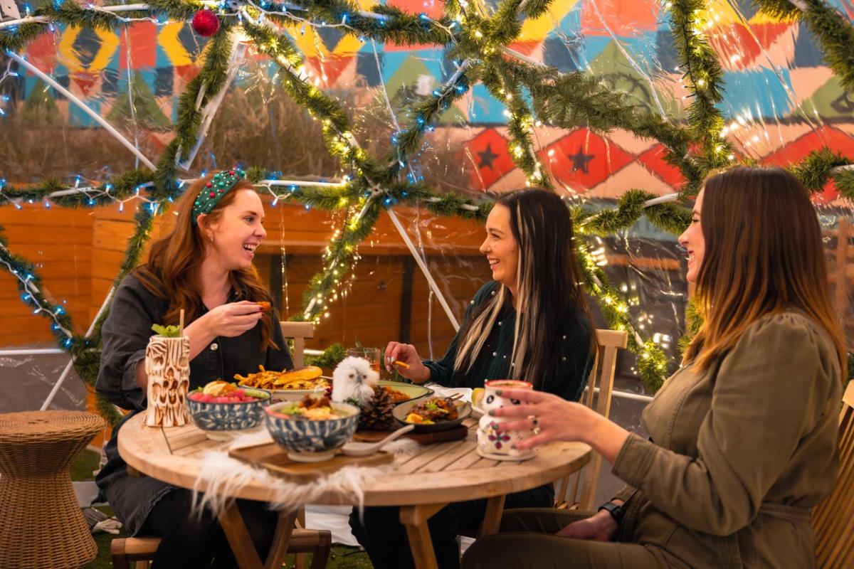 Three women enjoy a festive meal at a table adorned with colorful dishes, surrounded by twinkling lights and vibrant decorations.