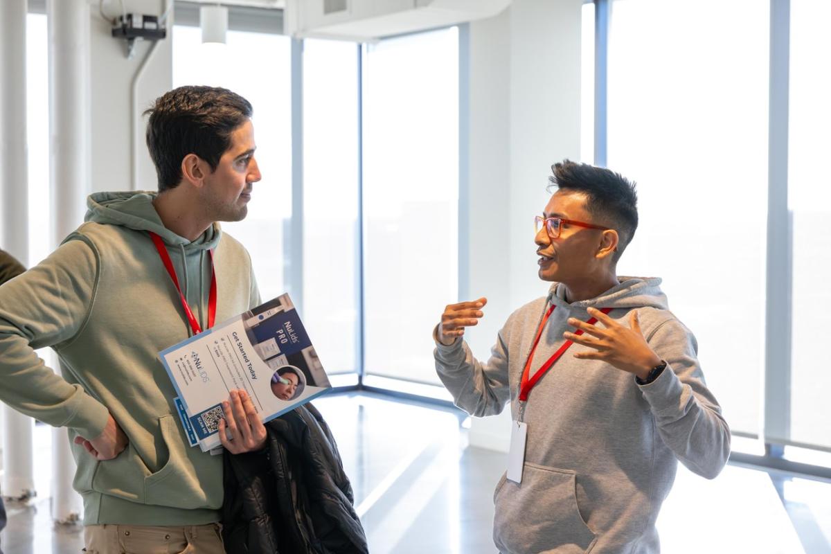 Two men engage in conversation indoors, with large windows providing a bright backdrop. One holds a brochure, while both wear name tags.