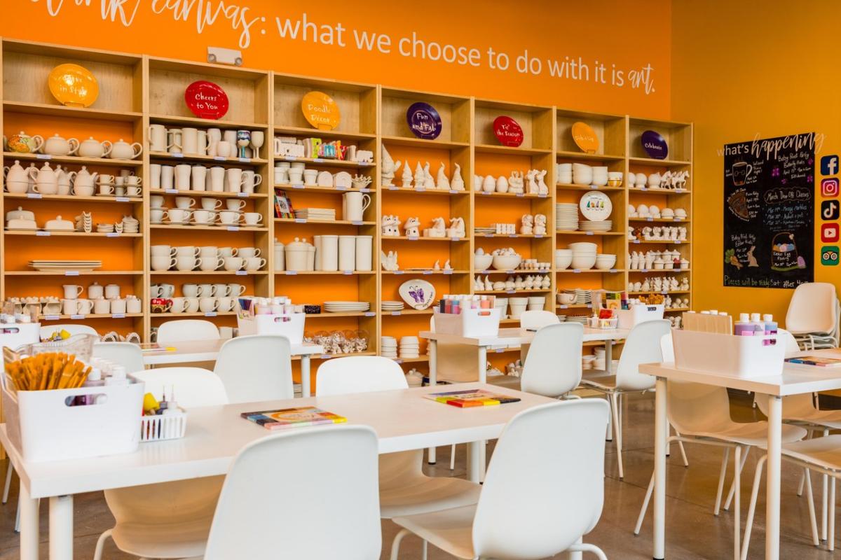 Brightly lit art studio featuring white tables and chairs, shelves filled with pottery and art supplies, and an orange accent wall with inspirational text.