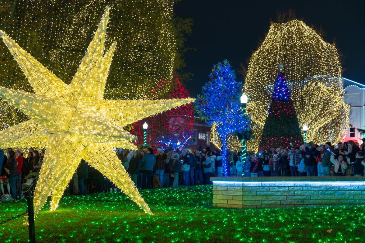 A vibrant display of holiday lights features a large star and decorated trees, with crowds enjoying the festive atmosphere at night at the Bentonville city square.