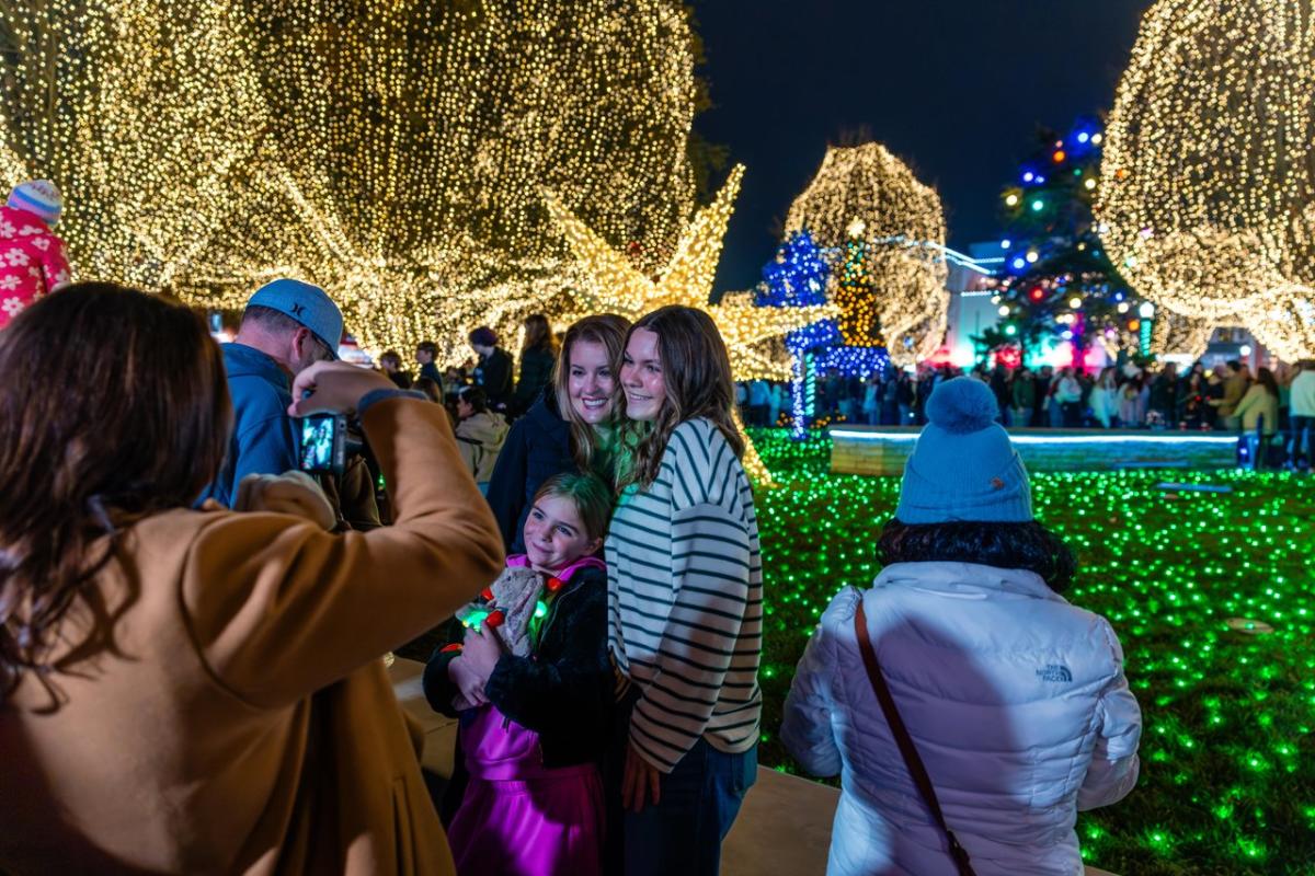Three people pose for a photo in a festive, illuminated park filled with colorful lights and decorated trees. A crowd enjoys the vibrant atmosphere of the Benotnville Square.
