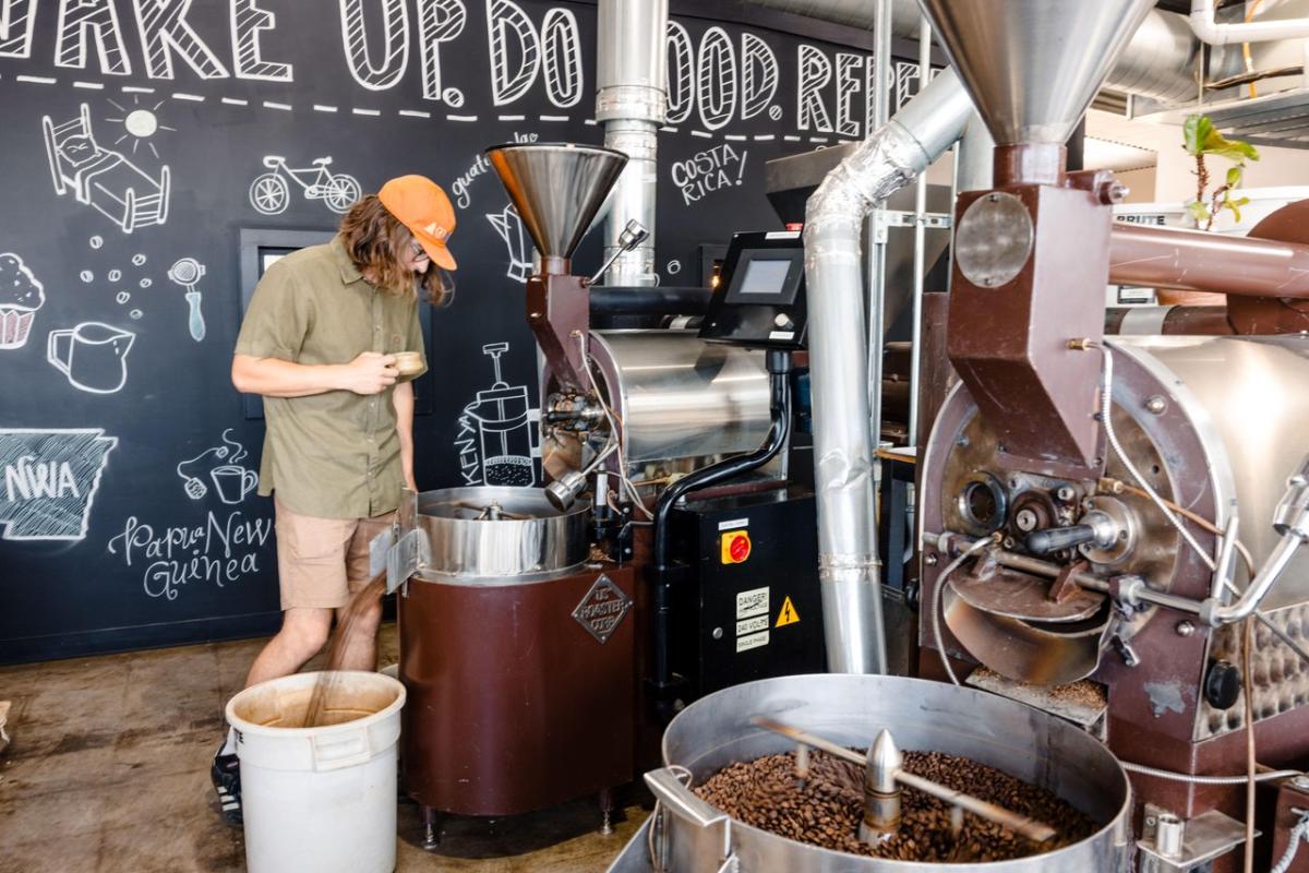 A barista operates a coffee roaster in a modern café, surrounded by equipment and a chalkboard wall with coffee-related illustrations.