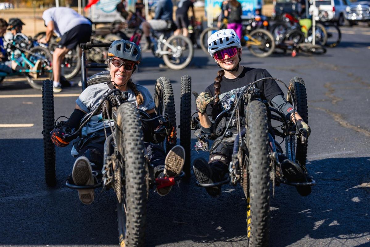 Two women smile while seated in handcycles, showcasing adaptive cycling. A busy outdoor event with various bikes and participants is in the background.