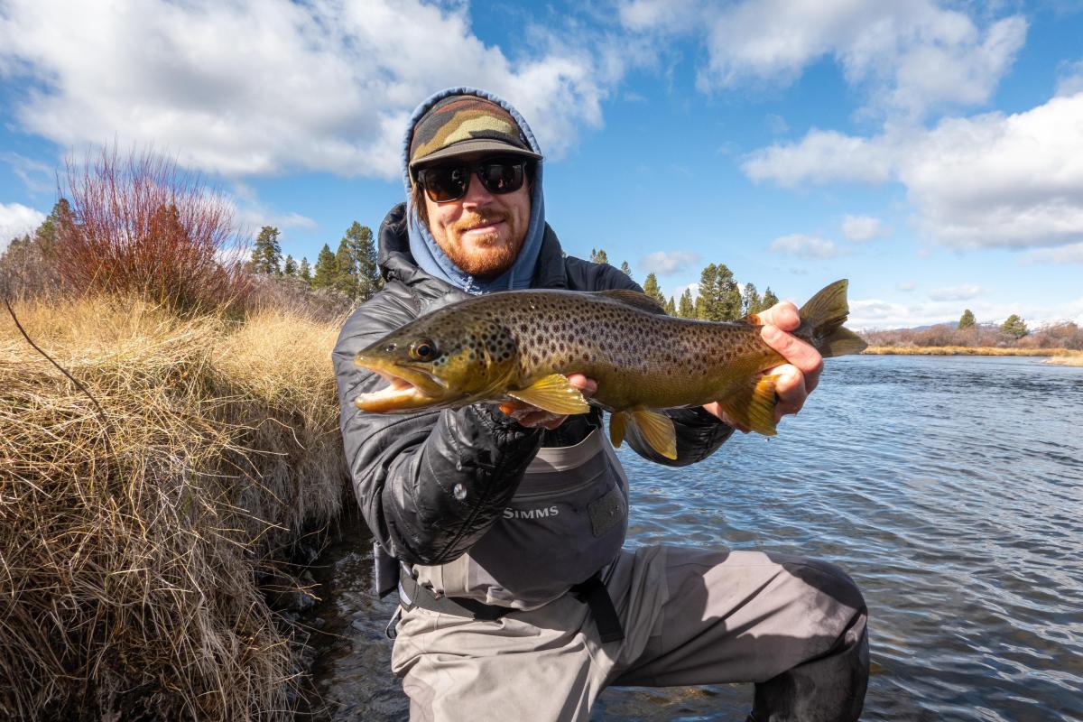 A person in fishing gear holds a large brown trout by a riverbank, surrounded by tall grass and trees under a blue sky with clouds.