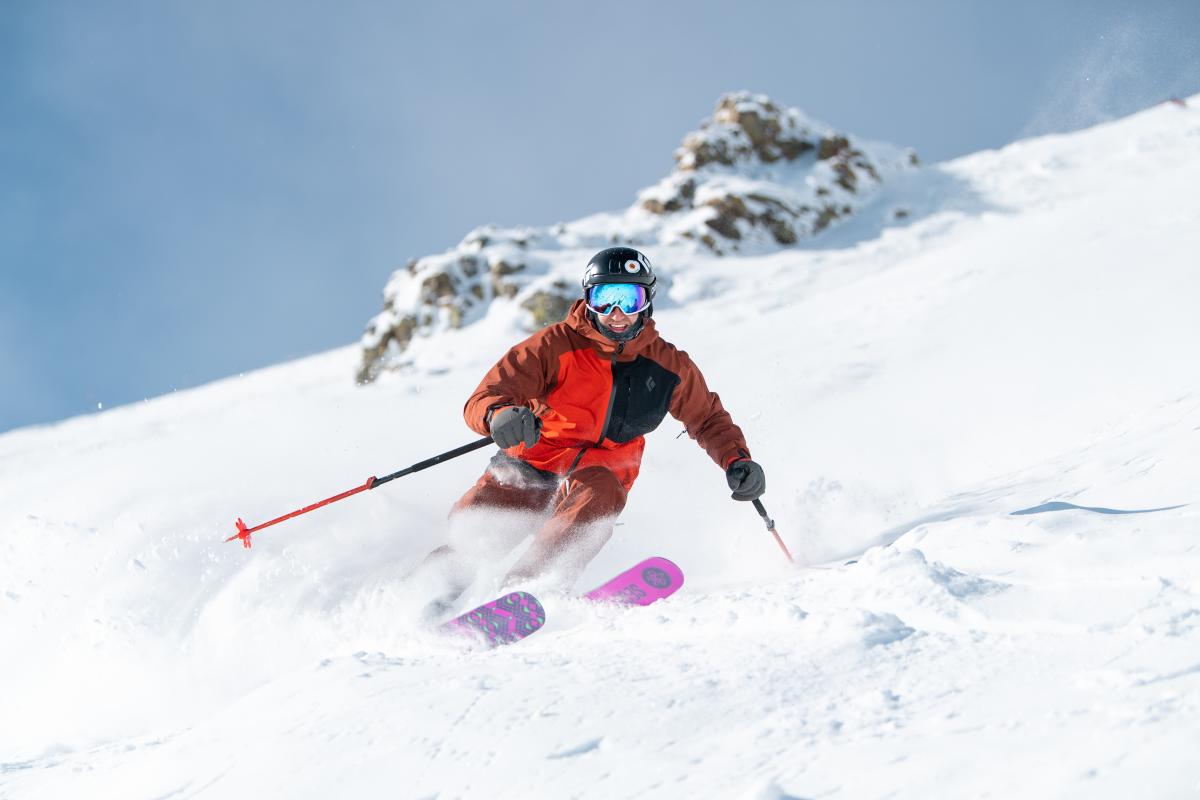 A skier in an orange jacket navigates a snowy slope, creating a spray of powder against a backdrop of rocky terrain and blue sky.