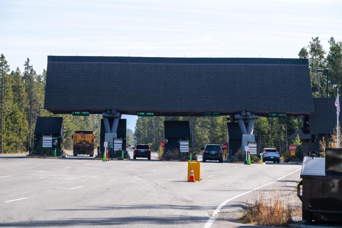 Entrance station with a large, wooden-roofed structure, flanked by trees and a clear blue sky. Road leads to the entry points.