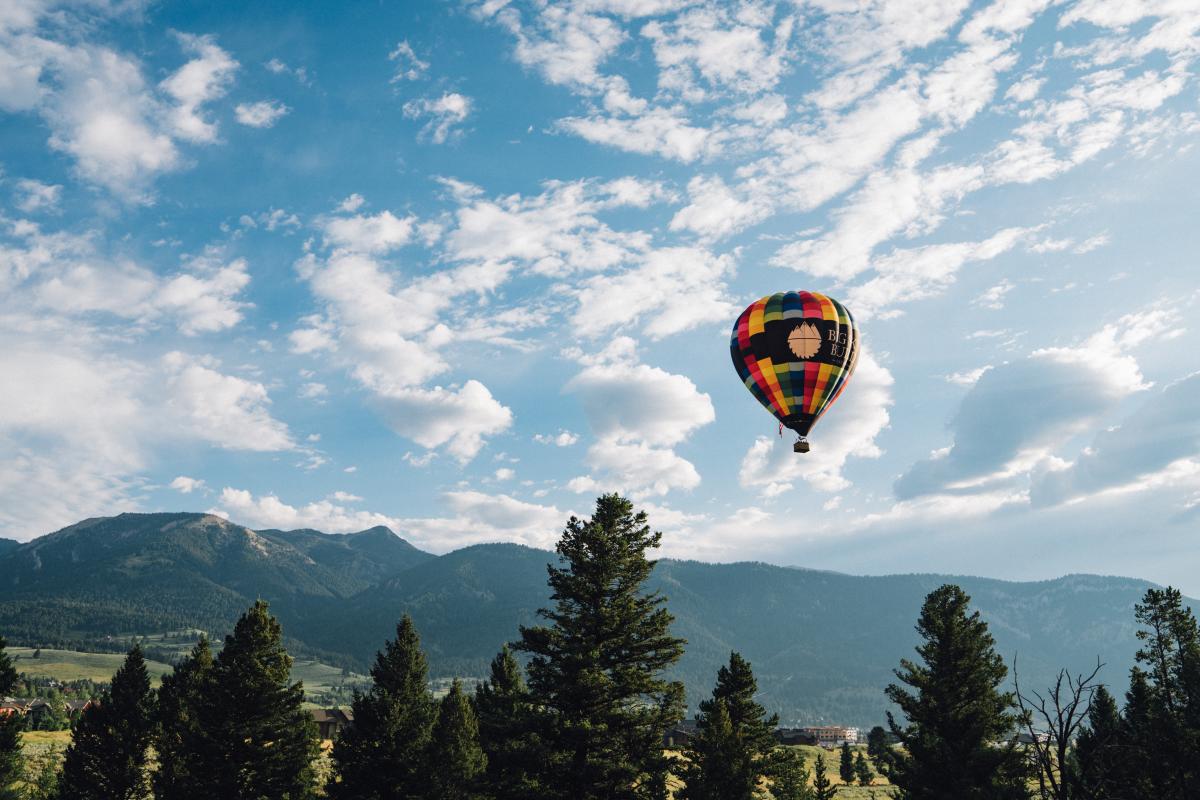 A colorful hot air balloon floats above lush green trees and rolling mountains under a bright blue sky with scattered clouds.