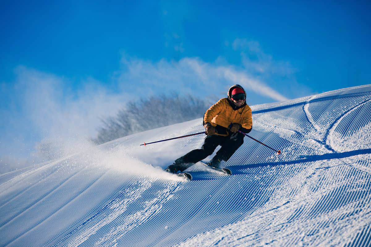 A skier is headed down a slope toward the camera with a yellow jacket and black pants on, two poles are pointed towards the ground where snow is spraying up.
