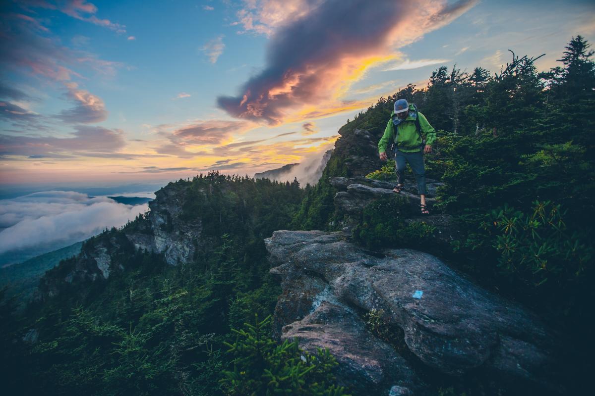 A man in a green jacket hikes over rocky terrain on the side of a mountain with clouds below.