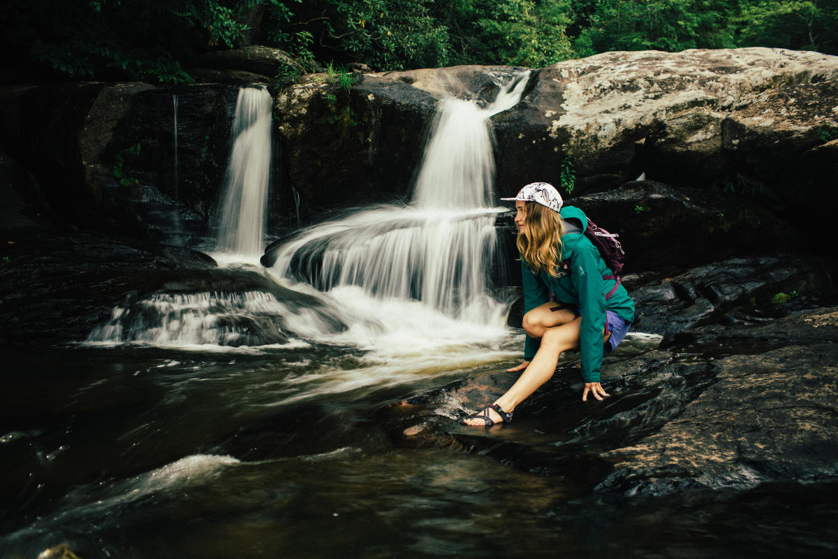 A female hiker crouches beside a small waterfall cascading over large boulders.