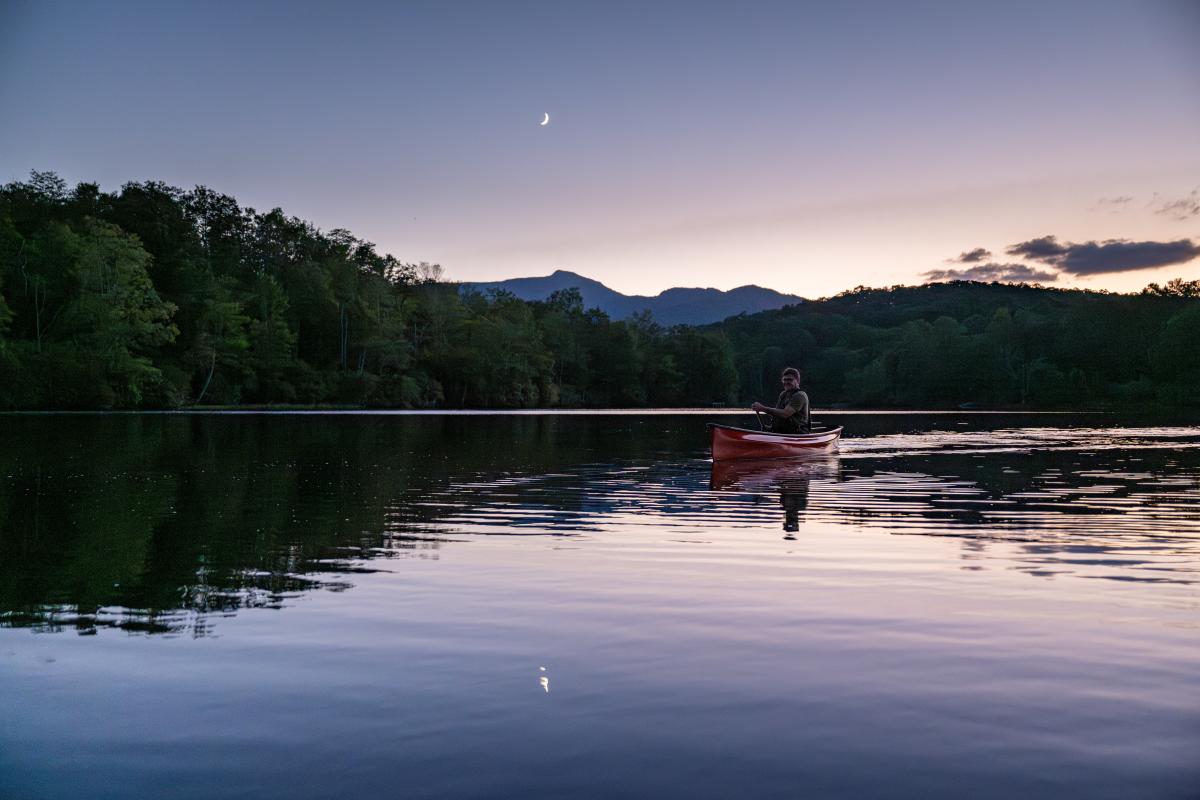 A paddler in a canoe glides across a calm mountain lake, the night sky illuminated purple by moonlight.