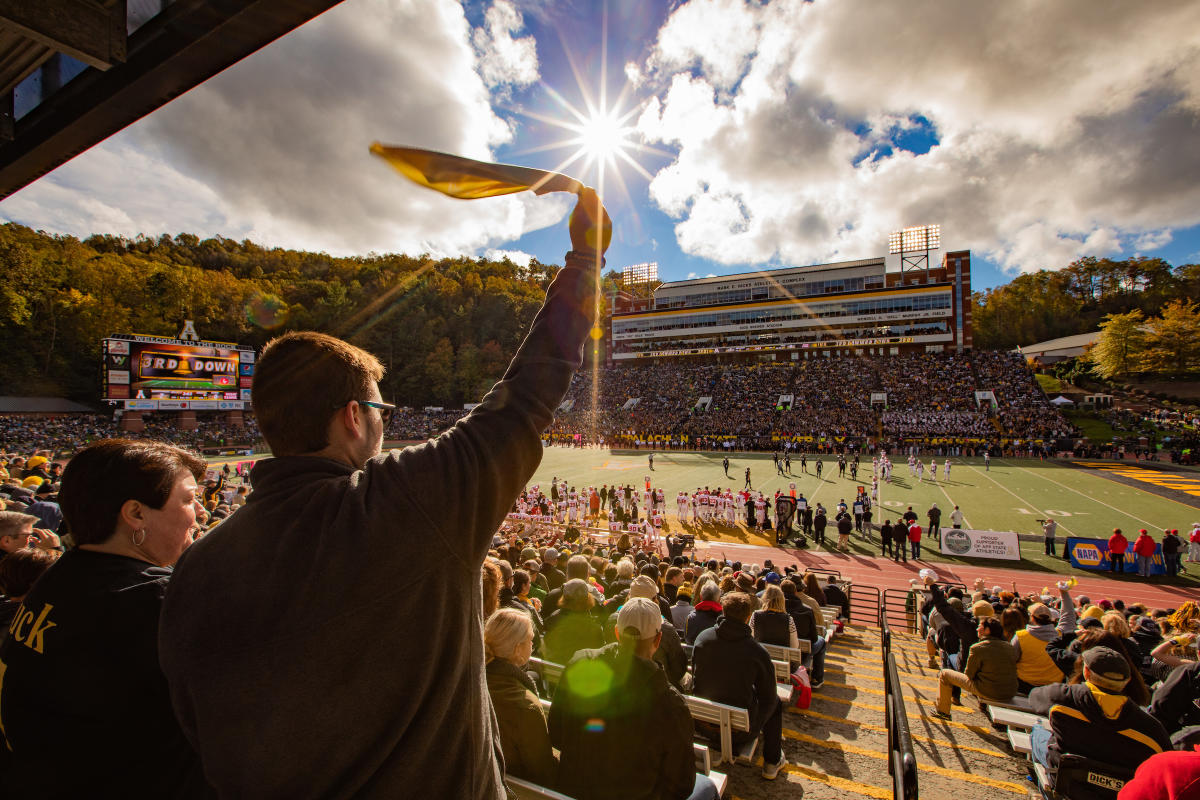 A man facing away is swinging a yellow towel over his head from bleachers. He is looking towards a football field with fans filling up the bleachers.