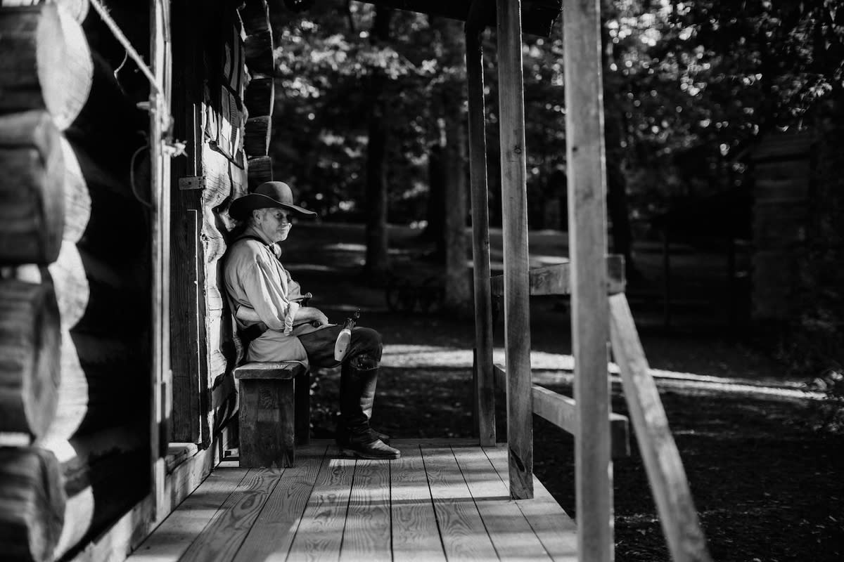 A man in colonial-era garb sits on the small porch of a historic log cabin, side-glancing at the viewer.