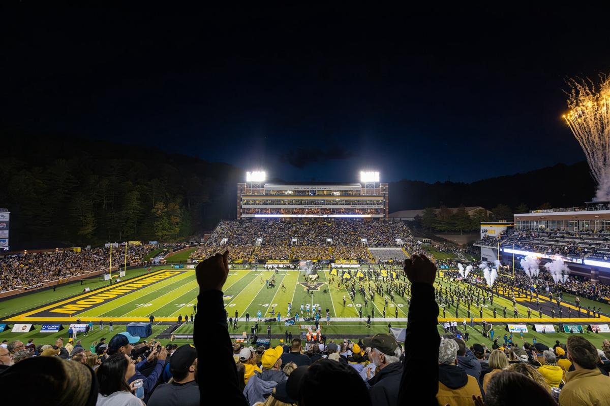 A fan raises two hands in the air as fireworks go off at a football game at night.