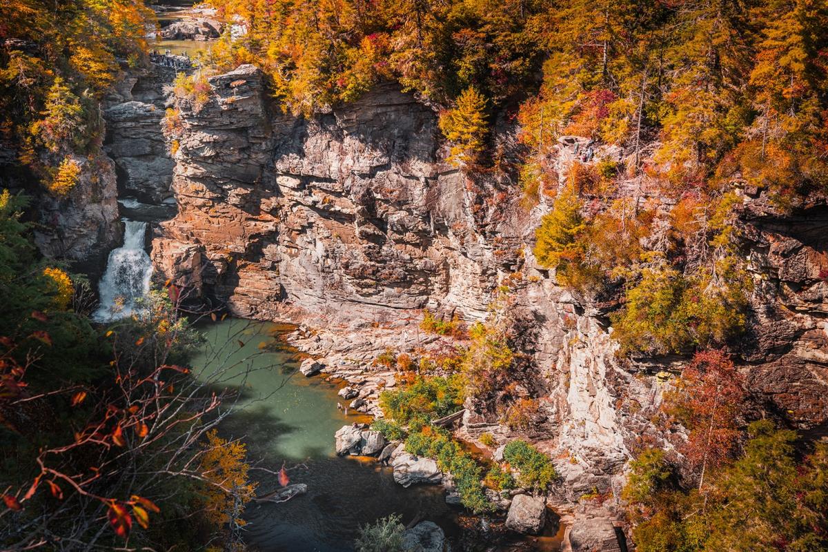 A large waterfall can be seen at the back of a canyon flowing into a river below. The top of the rocky ridge above the waterfall is lined with autumn-colored trees.