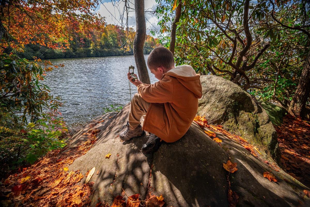 A child studies a compass while perched on a large boulder overlooking a scenic lake.