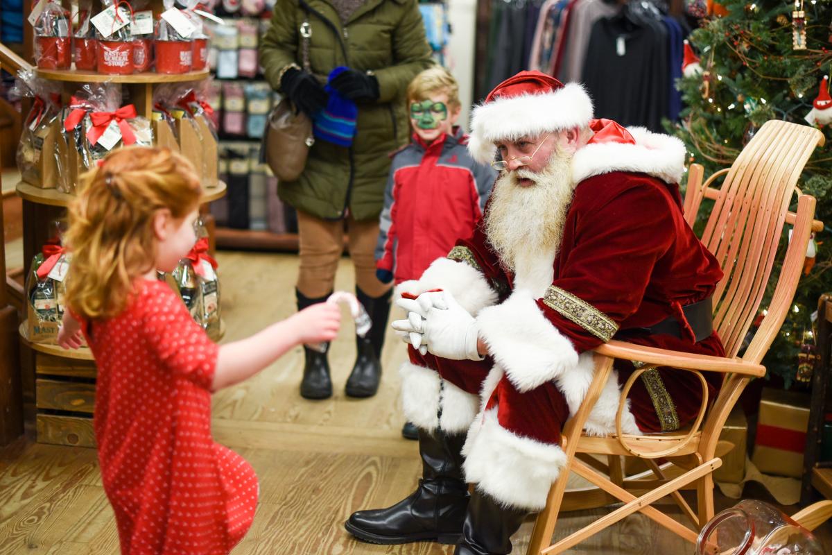 Santa Claus sits in a wooden rocking chair at Mast General Store, as a little girl in a red dress approaches with a candy cane.